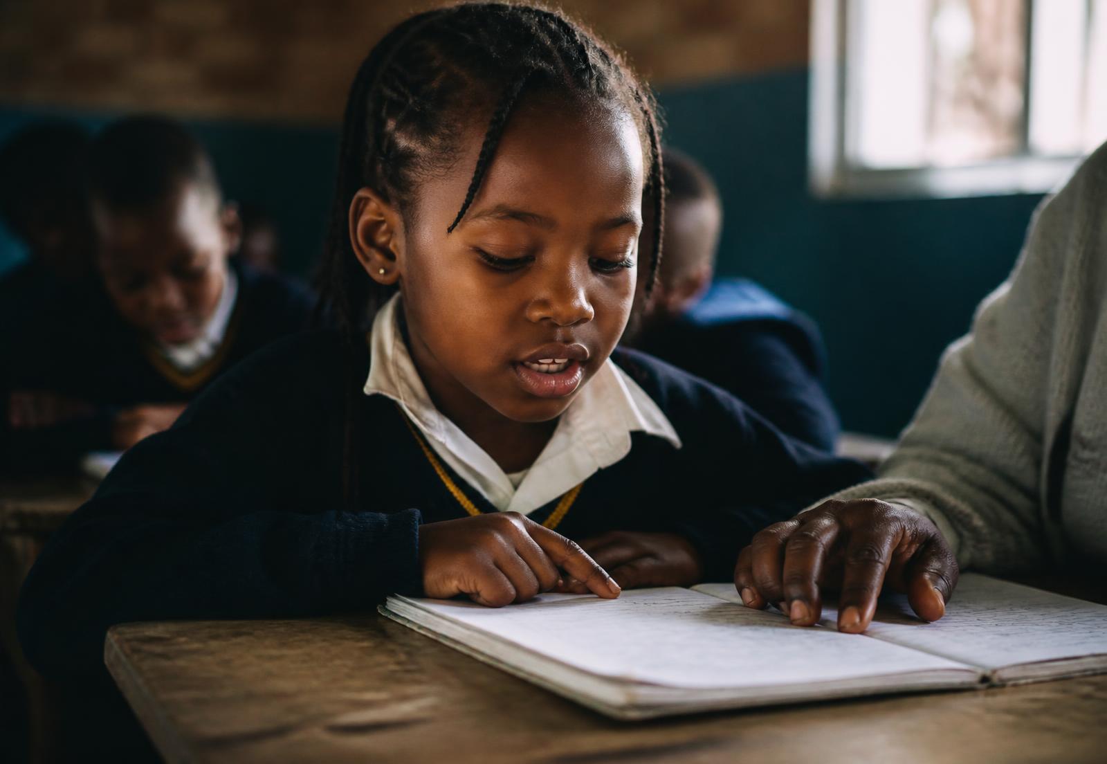 A young scholar reading aloud from an exercise book at a wooden desk, an adult's hand resting on the page beside her
