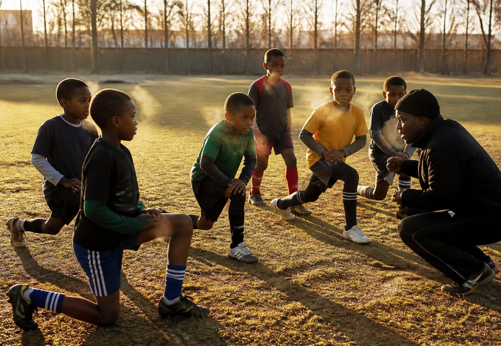 Children in mismatched sports kit doing warm-up drills on a grass field at sunrise while a coach crouches to give instruction